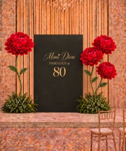 Black and gold birthday backdrop with custom gold lettering, oversized red flowers, tall green stems, and lush greenery arrangements for an elegant celebration.