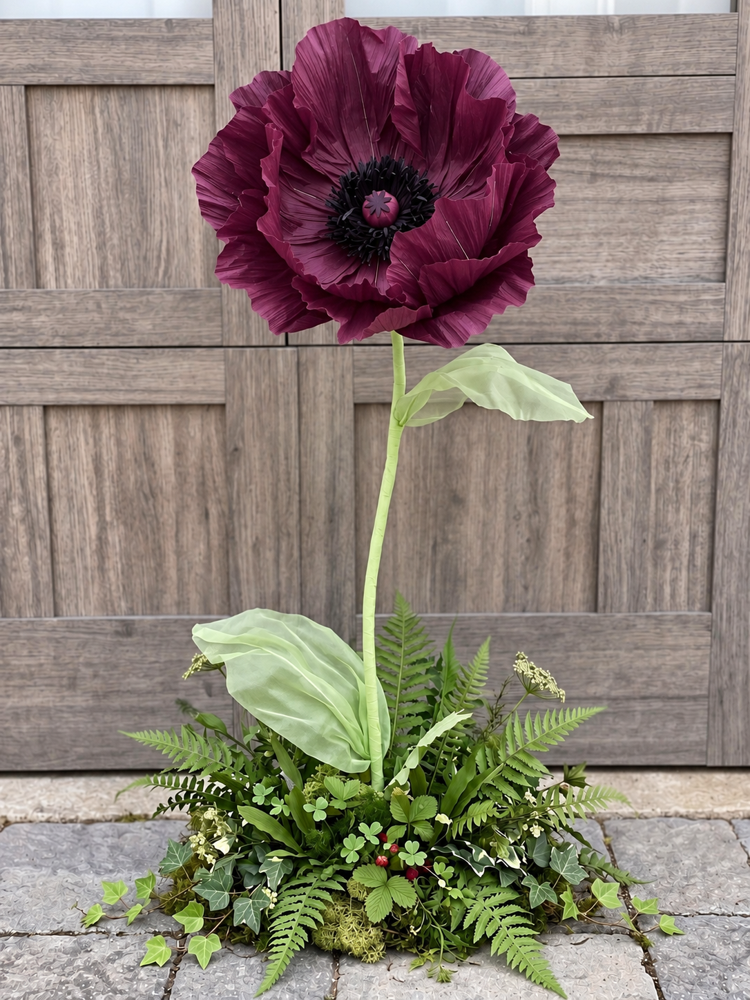 Giant burgundy poppy flower with a sculptural green stem, oversized leaves, and a lush greenery base with ferns, ivy, and moss.
