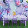 Mermaid and unicorn-themed birthday party setup featuring pastel pink, lavender and blue balloon garlands, iridescent sequin backdrop, seashell panel, white kids’ table, transparent purple ghost chairs, and magical themed decorations.