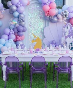 Mermaid and unicorn-themed birthday party setup featuring pastel pink, lavender and blue balloon garlands, iridescent sequin backdrop, seashell panel, white kids’ table, transparent purple ghost chairs, and magical themed decorations.