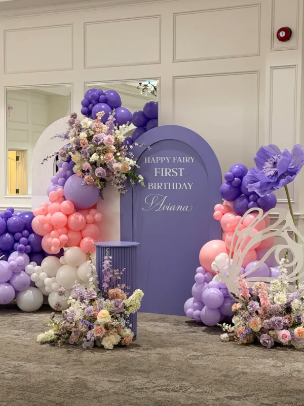 A custom pastel-themed first birthday backdrop setup at Montecassino Event Venue in Vaughan, featuring a purple arch with name lettering, layered balloon garlands, giant standing paper flowers, and lush floral arrangements.